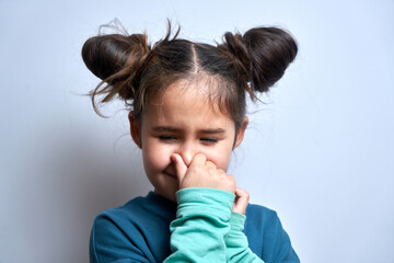 Little girl holding nose to avoid disgusted smell, pinches nose with fingers and holding breath isolated on white background - obrazy, fototapety, plakaty