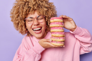 Overjoyed curly haired woman laughs positively holds pile of delicious sweet doughnuts enjoys...