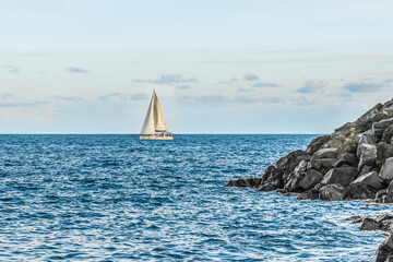 A white yacht sails past a pile of large boulders in blue sea water. Beautiful seascape