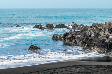 Fototapeta na wymiar Seascape on the Playa Maria Jimenez beach in Puerto de la Cruz, Spain. Volcanic rocks and black sand in blue water with white foam in the Canary Islands
