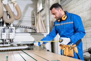 Handsome carpenter man in blue uniform use tape measure with machine at wood factory	