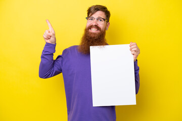 Redhead man with beard isolated on yellow background holding an empty placard and pointing side