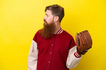 Redhead player man with beard with baseball glove isolated on yellow background looking to the side