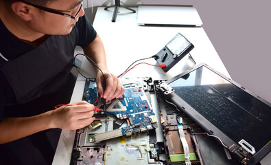 young man wearing glasses who is a technician computer technician A laptop motherboard repairman is using an IC meter to find faults on the motherboard for repair on his workbench.