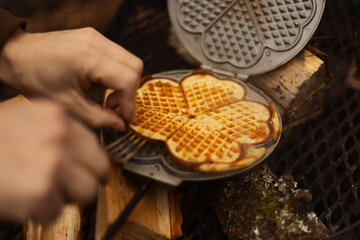 Hands holding a fork taking a waffle from a waffle iron on a campfire.