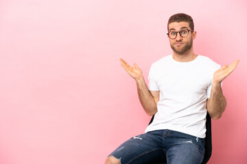 Young man sitting on a chair over isolated pink background having doubts while raising hands