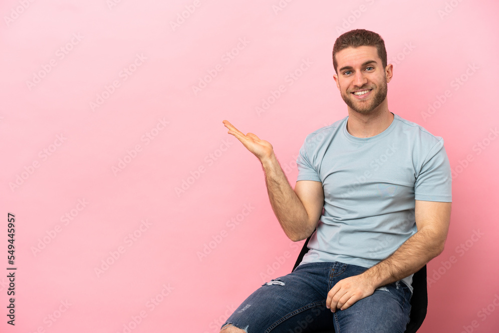 Poster Young man sitting on a chair over isolated pink background holding copyspace imaginary on the palm to insert an ad