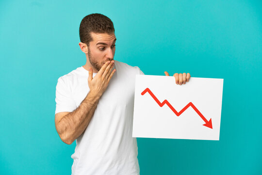 Handsome blonde man over isolated blue background holding a sign with a decreasing statistics arrow symbol with surprised expression