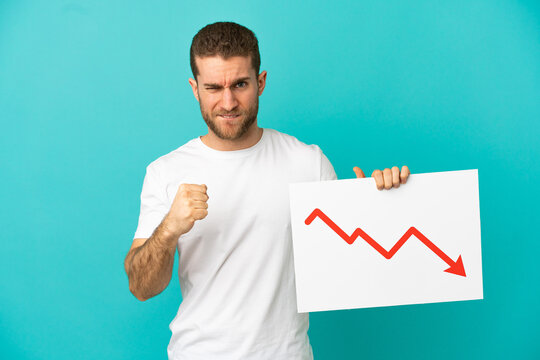 Handsome blonde man over isolated blue background holding a sign with a decreasing statistics arrow symbol and angry