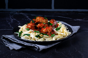 Meatballs in tomato sauce with fettuccine pasta served on black plate against dark background. Hot...