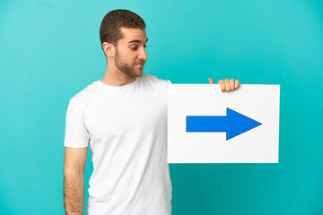 Handsome blonde man over isolated blue background holding a placard with arrow symbol