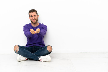 Young man sitting on the floor holding copyspace imaginary on the palm to insert an ad