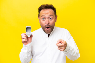 Middle age man holding a engagement ring isolated on yellow background surprised and pointing front