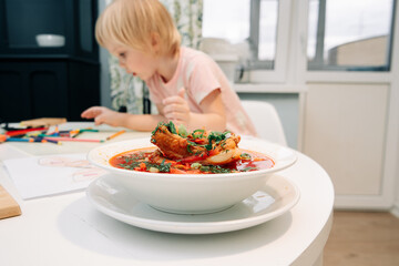 A plate of borscht on the table. A child draws with felt-tip pens in the background.