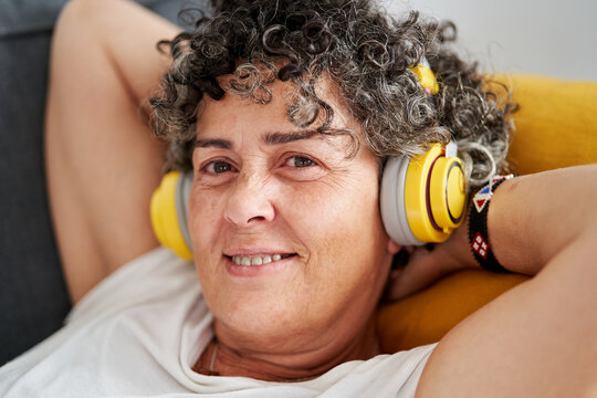 Mature Woman Wearing Wireless Headphones Lying On Couch With Hands Behind Head Looking At Camera