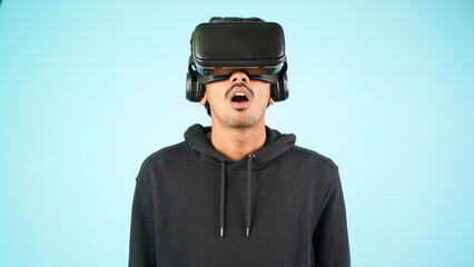 Young Indian Asian man using VR headset, glasses or goggles, isolated over blue colour indoor studio background, shocked surprised or amazed concept