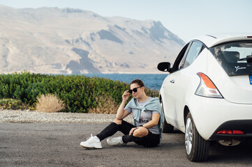 A beautiful brunette in sunglasses sits by a white car against the backdrop of the Mediterranean Sea and mountains