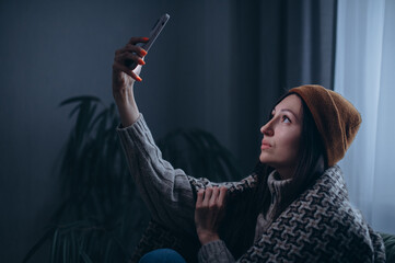 A young woman is trying to catch a connection during a power outage. The female sits on a couch in a dark room with no electricity, no communication, and no heating.