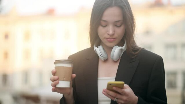 Successful young beautiful woman standing on the old street using smartphone and drinking coffee. Girl work on smart phone. Social networks, online shopping concept. Attractive business gir