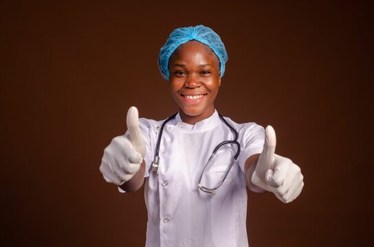 Smiling Nigerian female medic with a stethoscope showing thumb up gesture on a brown background