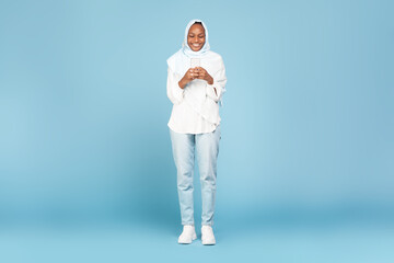 Full length shot of happy black muslim woman using smartphone, checking mobile application, blue background