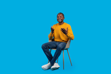 Relaxed black man sitting on chair and gesturing on blue