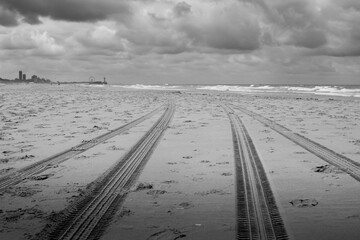 Tire tracks in the sand of a beach along the sea with a fierce sky and a Ferris wheel in the distance.