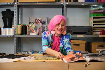 Seamstress with pink hair and colorfull clothes taking ideas from fashion magazines in a sewing workshop.