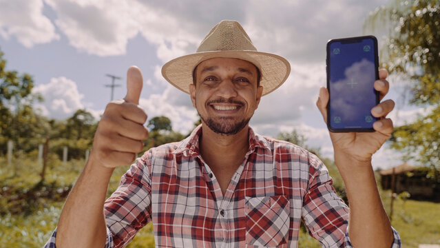 Young man farmer in hat smiling to camera and showing smartphone with chroma key screen. Latin happy male shepherd holding cellphone and demonstrating advertisement