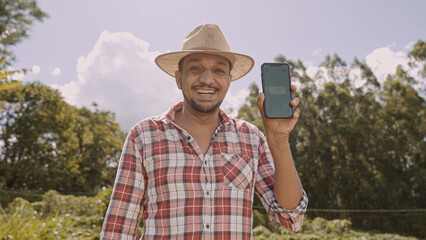 Young man farmer in hat smiling to camera and showing smartphone with chroma key screen. Latin...