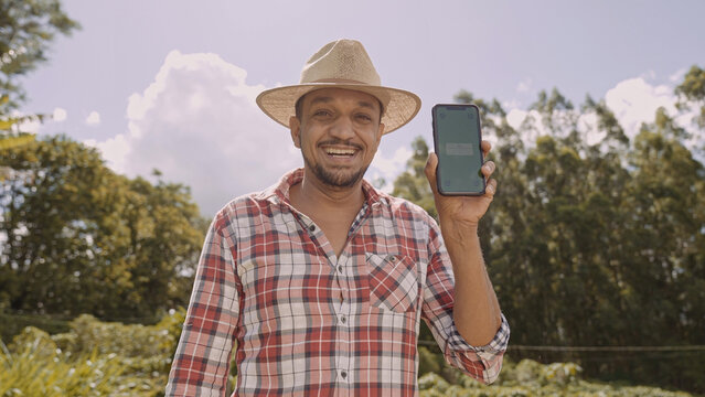 Young man farmer in hat smiling to camera and showing smartphone with chroma key screen. Latin happy male shepherd holding cellphone and demonstrating advertisement