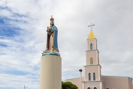 Imagem de Nossa senhora em primeiro plano e detalhe da Paróquia Nossa Senhora D'Abadia ao fundo, em um dia de céu nublado. Igreja na cidade de Anápolis em Goiás.