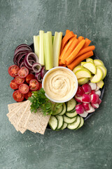 Healthy snack chickpea hummus with various fresh vegetables and cereals on a gray platter. Cucumbers, radishes, celery, carrots, tomatoes, zucchini, red onions, parsley, cilantro. Healthy food