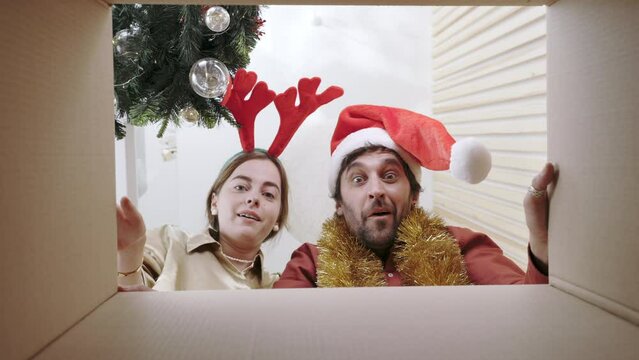 Caucasian family with shocked facial expression looking in carton box and putting hands inside. Happy couple in santa hats unpacking christmas presents at home. Unboxing gift. Holiday