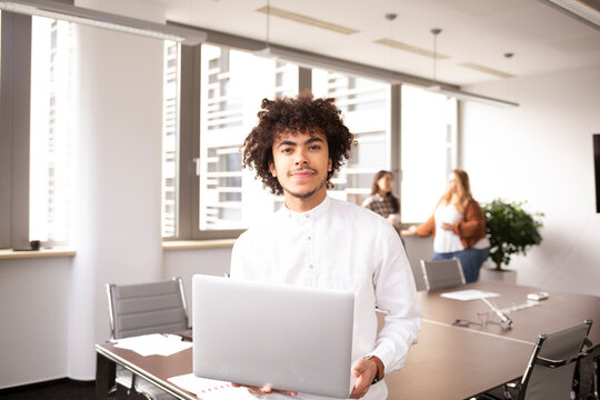 Young man in the office, in foreground looking at the camera with his laptop