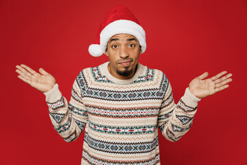 Merry young man wear Christmas sweater Santa hat posing spread hands shrugging shoulders looking puzzled, have no idea isolated on plain red background Happy New Year 2023 celebration holiday concept