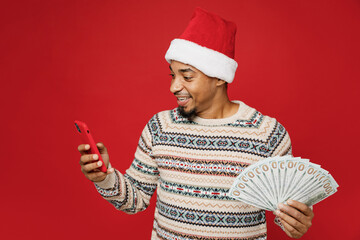 Merry smiling young man wear Christmas sweater Santa hat posing use mobile cell phone hold fan of...