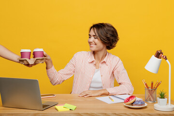 Young smiling employee business woman wearing shirt take takeaway paper cup coffee to go sit work at office desk with pc laptop isolated on plain yellow color background. Achievement career concept.