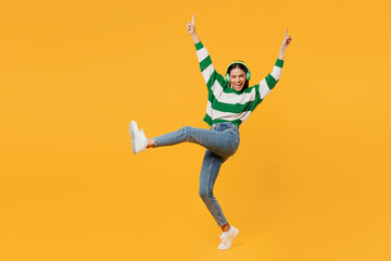 Full body cheerful happy young latin woman in casual cozy green knitted sweater headphones raise up hands listen to music isolated on plain yellow background studio portrait People lifestyle concept