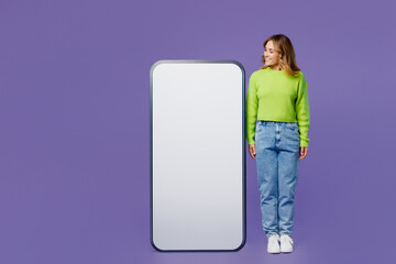 Full body smiling happy fun young woman 30s she wearing casual green knitted sweater stand look at big huge blank screen mobile cell phone with area isolated on plain pastel purple background studio.