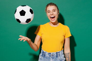 Young cool happy woman fan wear basic yellow t-shirt cheer up support football sport team hold in hand toss up soccer ball look camera watch tv live stream isolated on dark green background studio.