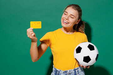 Young happy woman fan in basic yellow t-shirt cheer up support football sport team hold soccer ball yellow card propose player retire from field watch tv live stream isolated on dark green background.