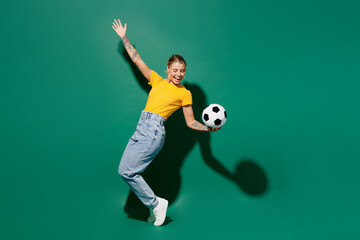 Fototapeta na wymiar Full body fun young woman fan wear yellow t-shirt cheer up support football sport team hold soccer ball watch tv live stream stand on toes leaning back raise up hand isolated on dark green background.
