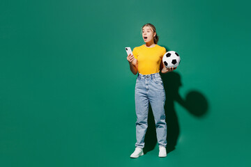 Fototapeta na wymiar Full body young surprised woman fan wear basic yellow t-shirt cheer up support football sport team hold soccer ball watch tv live stream use mobile cell phone isolated on dark green background studio