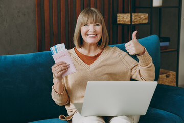 Traveler elderly woman 50s year old wearing sweater hold passport ticket sit on sofa stay at home flat spend free spare time in living room Tourist travel abroad rest getaway. Air flight trip concept.
