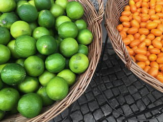 A straw basket with fresh kumquats and limes on street pavers closeup.