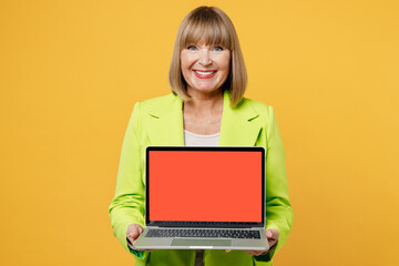 Naklejka premium Elderly smiling happy IT woman 50s years old wear green jacket white t-shirt hold use work on laptop pc computer with blank screen workspace area isolated on plain yellow background studio portrait.