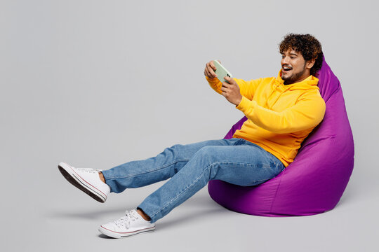 Full body young Indian man he wear casual yellow hoody sit in bag chair using play racing app on mobile cell phone hold gadget smartphone for pc video games isolated on plain grey background studio
