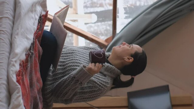 Vertical video. Young woman reading book and drinking hot tea or chocolate while sitting on the bed during winter vacation in geo dome glamping tent 