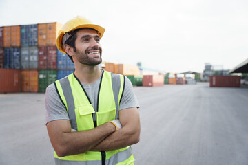 Portrait of positive Caucasian man worker smiling wearing safety vest and hard hat standing in front of container yard at the background. Optimistic man working on site.